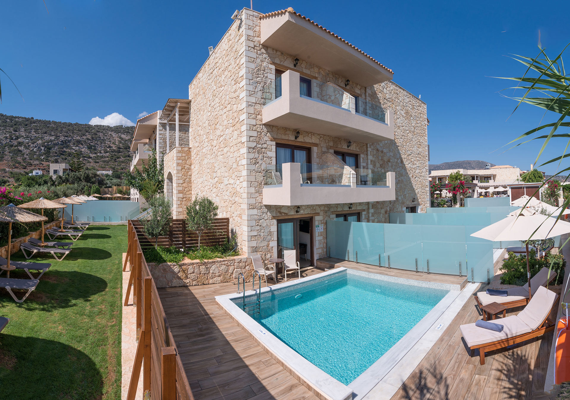 Εξωτερική όψη πετρόχτισου ξενοδοχείου και πισίνας που έχει διαχωριστίκο τζάμι με θεα τον ουρανό με το βουνό και με γρασίδι και σετ ξαπλώστρες