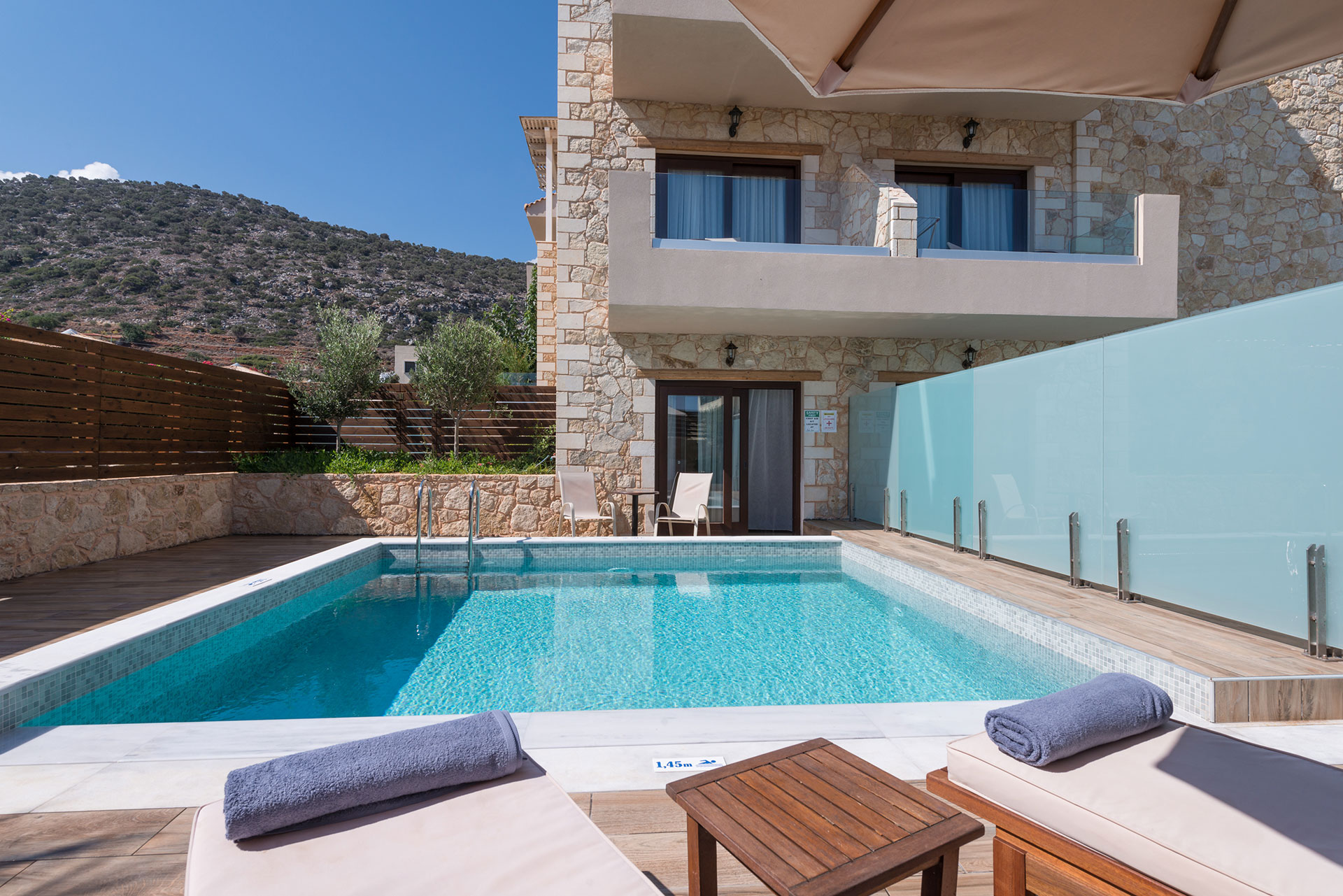 Εξωτερική όψη πετρόχτισου ξενοδοχείου και πισίνας που έχει διαχωριστίκο τζάμι και θέα τον ουρανό με το βουνό 